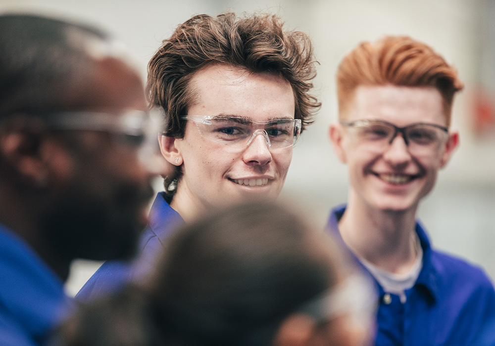 A young man, who is wearing protective eyewear, smiles as he enjoys his engineering class with peers - they are all wearing blue coveralls.